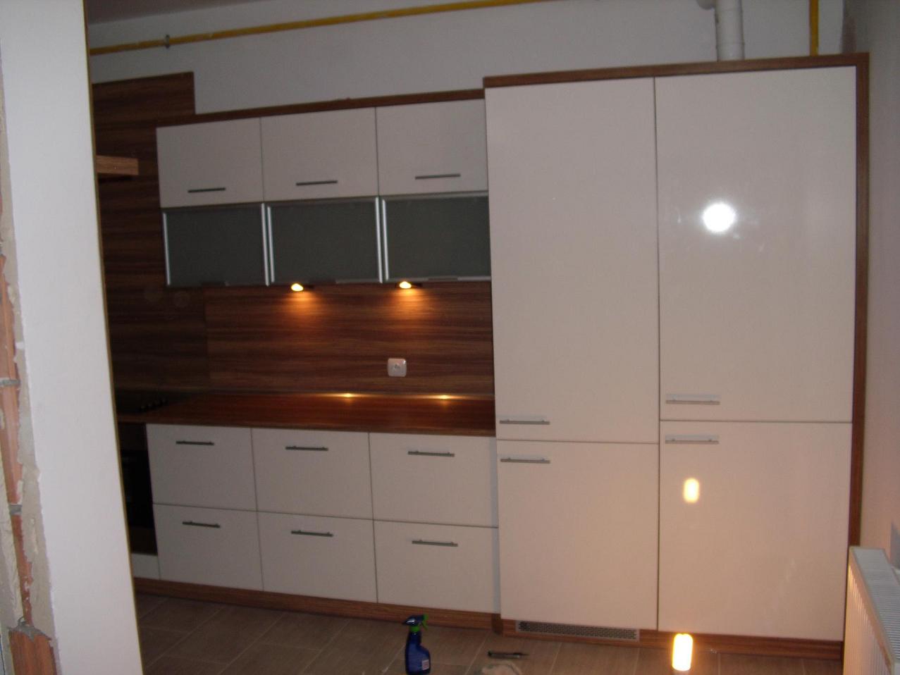 Nowoczesna zabudowa kuchenna z białymi, błyszczącymi frontami i drewnianymi akcentami, podświetlana punktowo, zrealizowana w Buk. Widoczny fragment ściany w trakcie remontu.